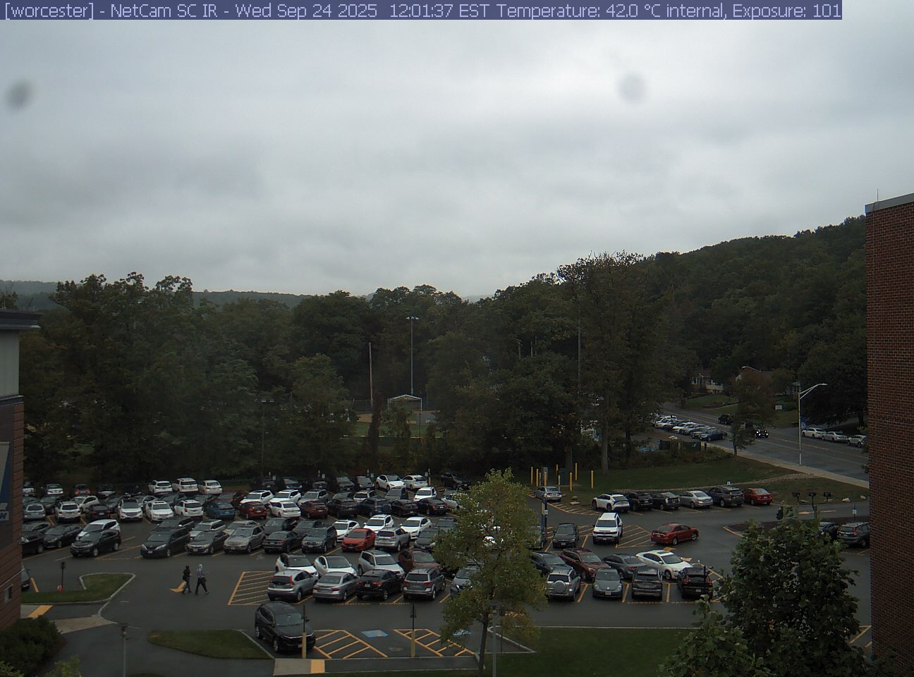 A wooded hill rises beyond a campus car park and a ball field, with a brick wall at the right edge. Cars, buildings and sky take up much of the view.