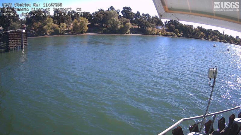 Yellow, muted orange and green trees line the opposite bank and reflect in the Sacramento River. A roof edge, railing and metal arm appear at the right of the wide stretch of water.