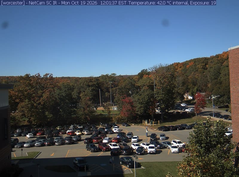 A wooded hill rises beyond a campus car park and a ball field, with a brick wall at the right edge. Cars, buildings and sky take up much of the view.