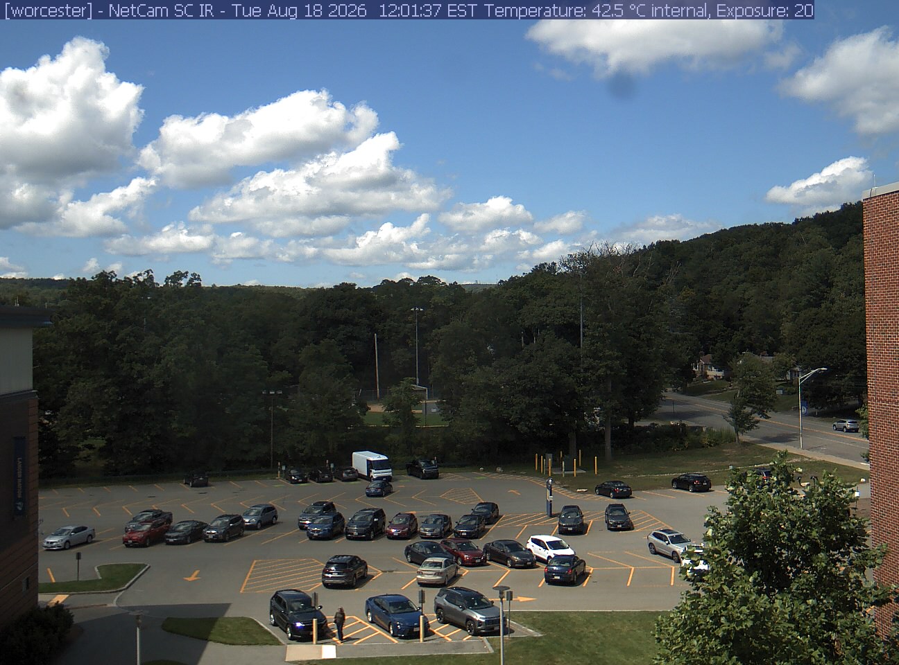 A wooded hill rises beyond a campus car park and a ball field, with a brick wall at the right edge. Cars, buildings and sky take up much of the view.
