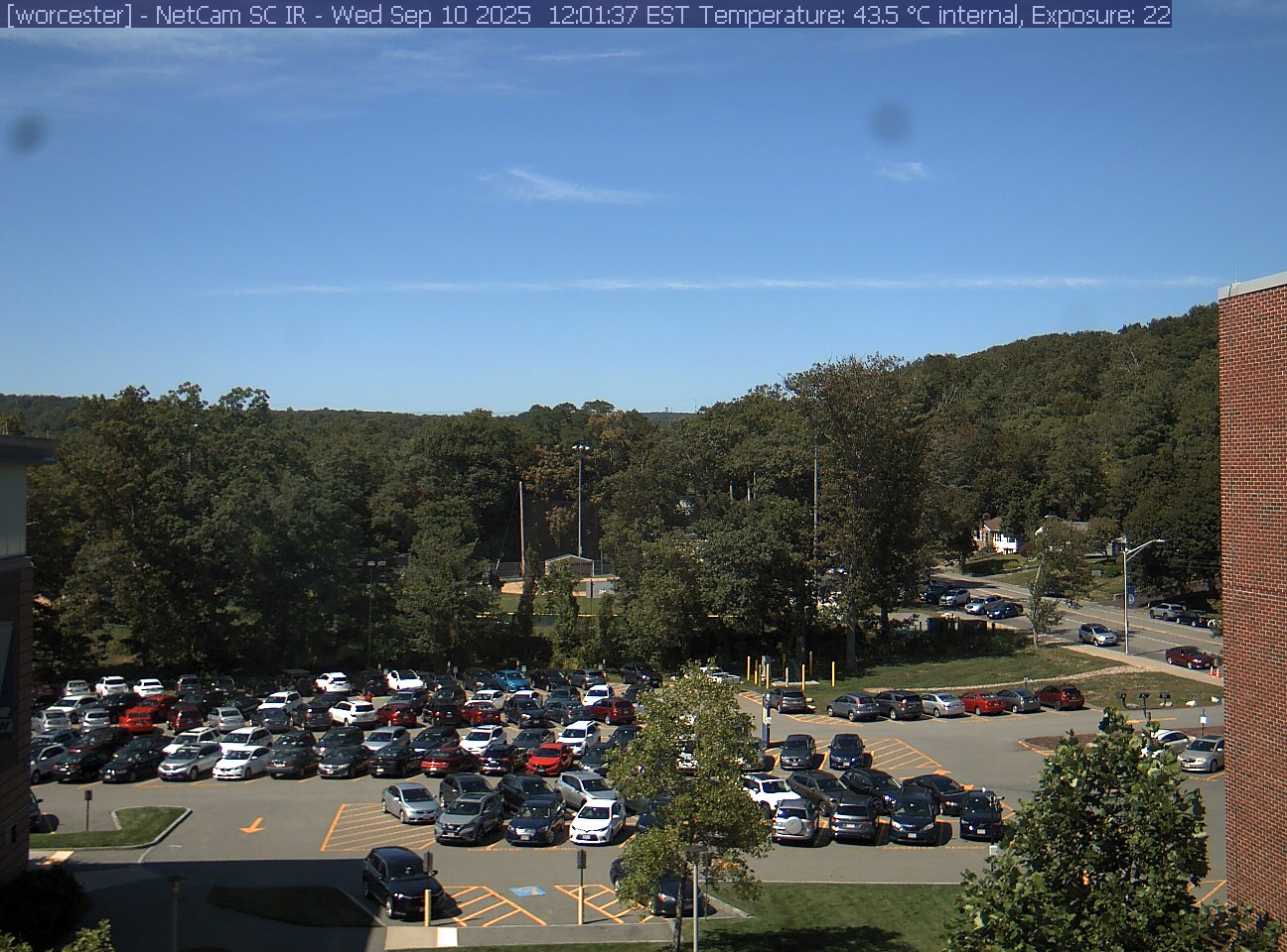 A wooded hill rises beyond a campus car park and a ball field, with a brick wall at the right edge. Cars, buildings and sky take up much of the view.