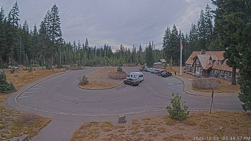 A stone-and-shingle park building beside a circle of parking, ringed by tall firs and pines.