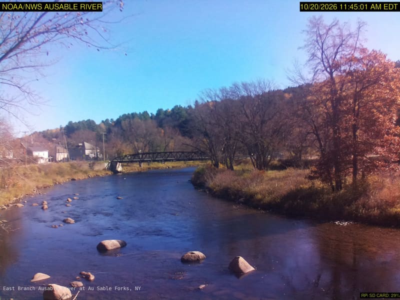 A broad rocky river with a footbridge and large yellow, orange, red and green tree crowns along both banks. The original credits NOAA/NWS; its clock is 15 minutes earlier than the USGS listing, which is a delivery timestamp here.