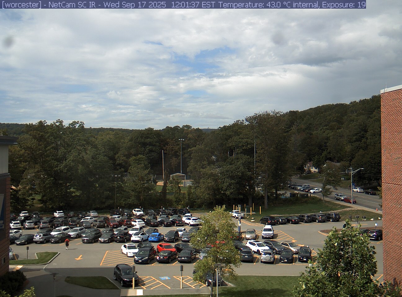 A wooded hill rises beyond a campus car park and a ball field, with a brick wall at the right edge. Cars, buildings and sky take up much of the view.