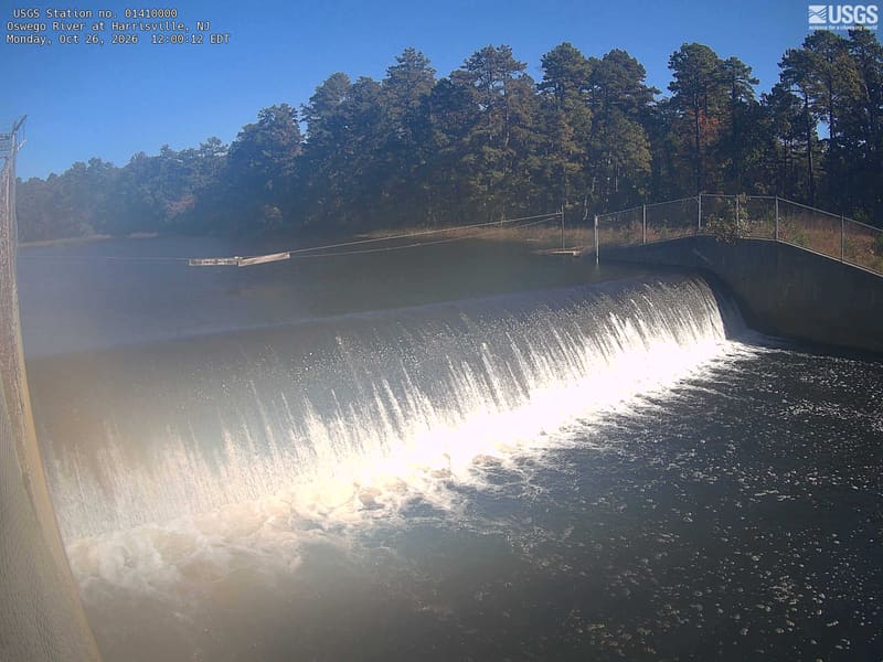 Wide sparkling spillway with lake above it and dense pines along the far shore, with scattered muted red deciduous foliage at left.