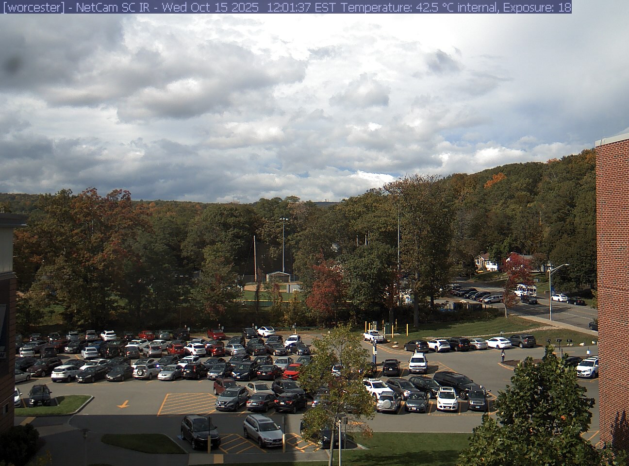 A wooded hill rises beyond a campus car park and a ball field, with a brick wall at the right edge. Cars, buildings and sky take up much of the view.