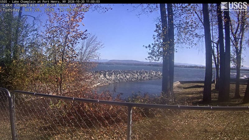 Yellowing trees and a red shrub frame a lake and distant low mountains, with large trunks and a waterfront fence in the foreground. The center has a little haze.