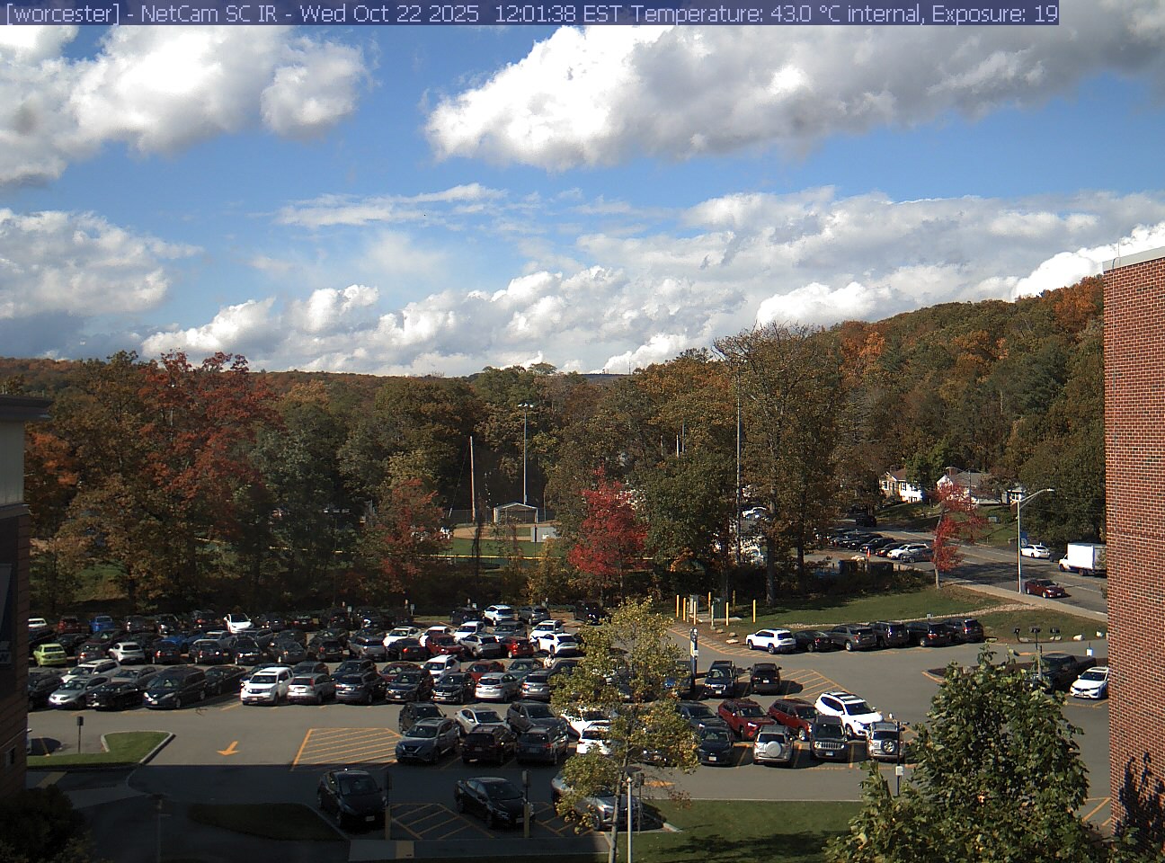 A wooded hill rises beyond a campus car park and a ball field, with a brick wall at the right edge. Cars, buildings and sky take up much of the view.