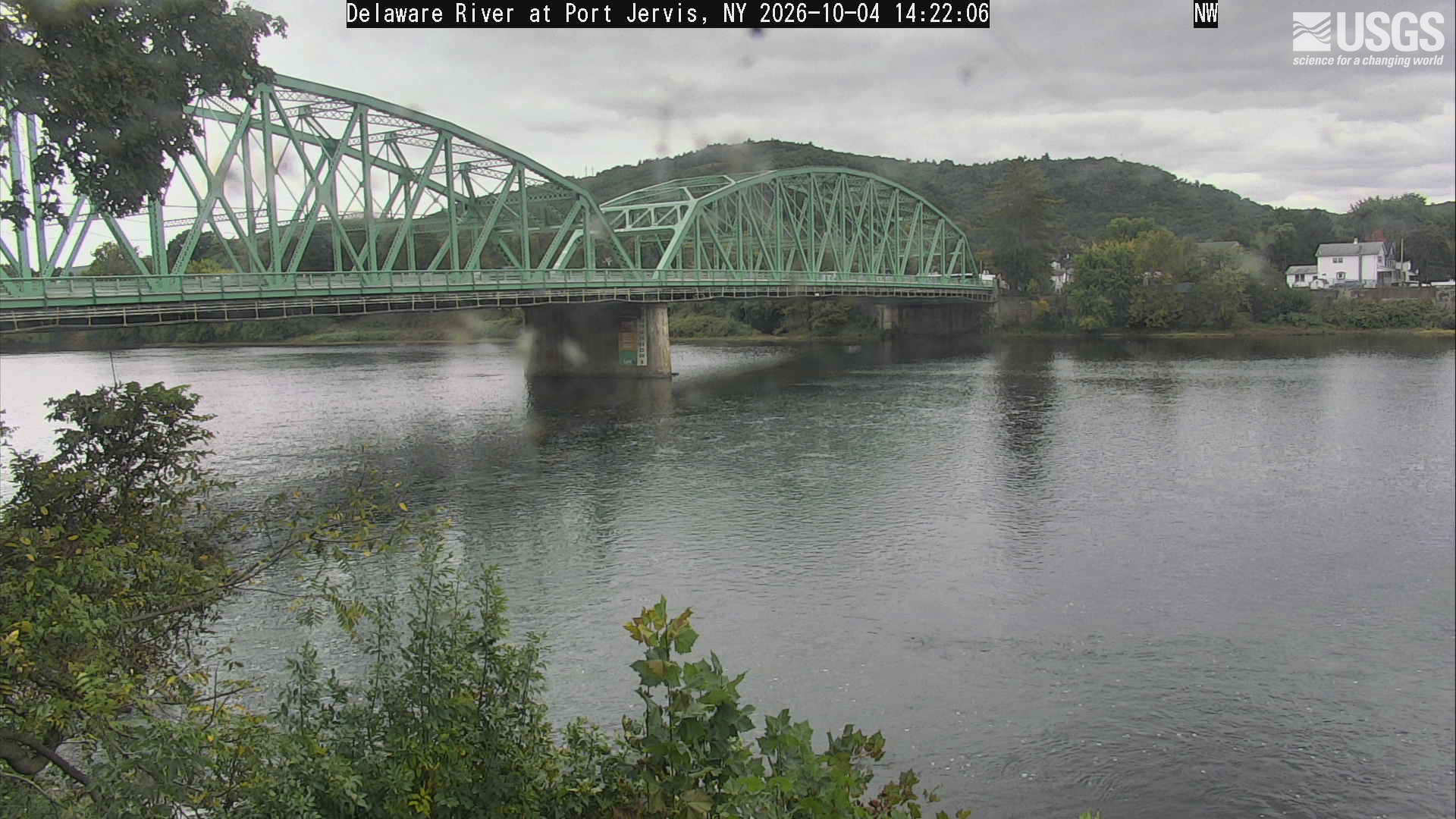 A wide river and green metal bridge below a wooded hill, with near green branches and small yellow patches on the opposite bank. Bridge and water dominate, and lens spots soften portions of the scene.