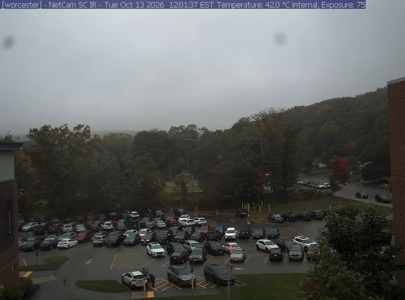 A wooded hill rises beyond a campus car park and a ball field, with a brick wall at the right edge. Cars, buildings and sky take up much of the view.