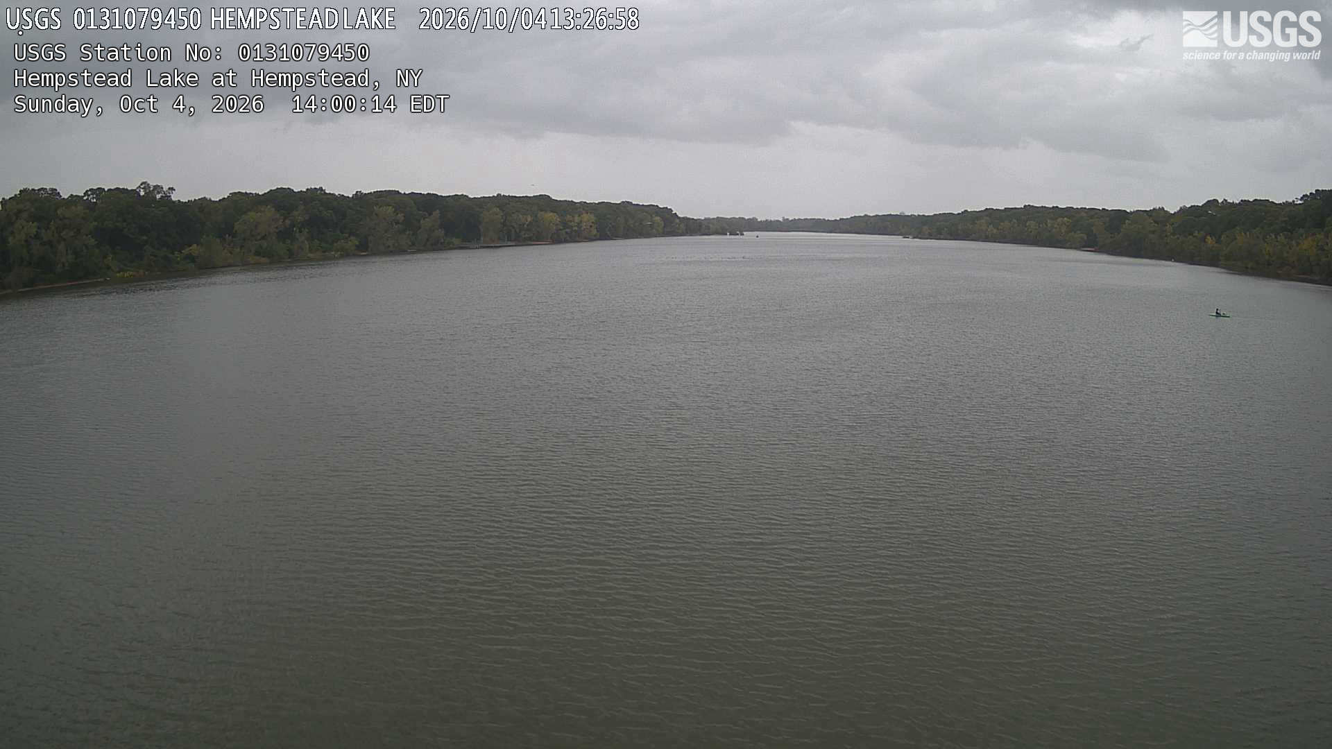 A wide calm lake beneath gray clouds, edged by a continuous low wooded bank. The native clock precedes the service overlay by about 33 minutes, with an uncontradicted October 4 date.