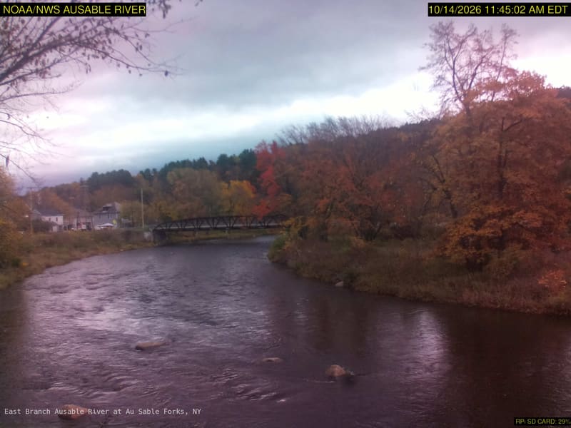 A broad rocky river with a footbridge and large yellow, orange, red and green tree crowns along both banks. Some softness remains, but broad crown color and the riverside setting are clear. The original credits NOAA/NWS; its clock is 15 minutes earlier than the USGS listing, which is a delivery timestamp here.