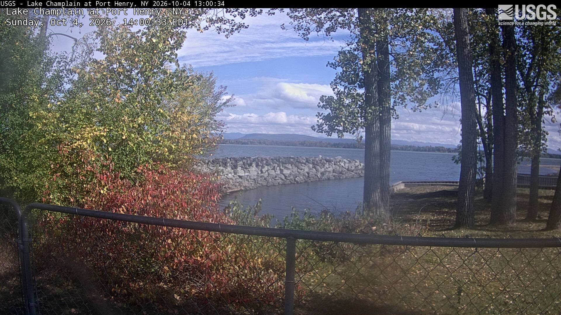 Yellowing trees and a red shrub frame a lake and distant low mountains, with large trunks and a waterfront fence in the foreground. The center has a little haze.