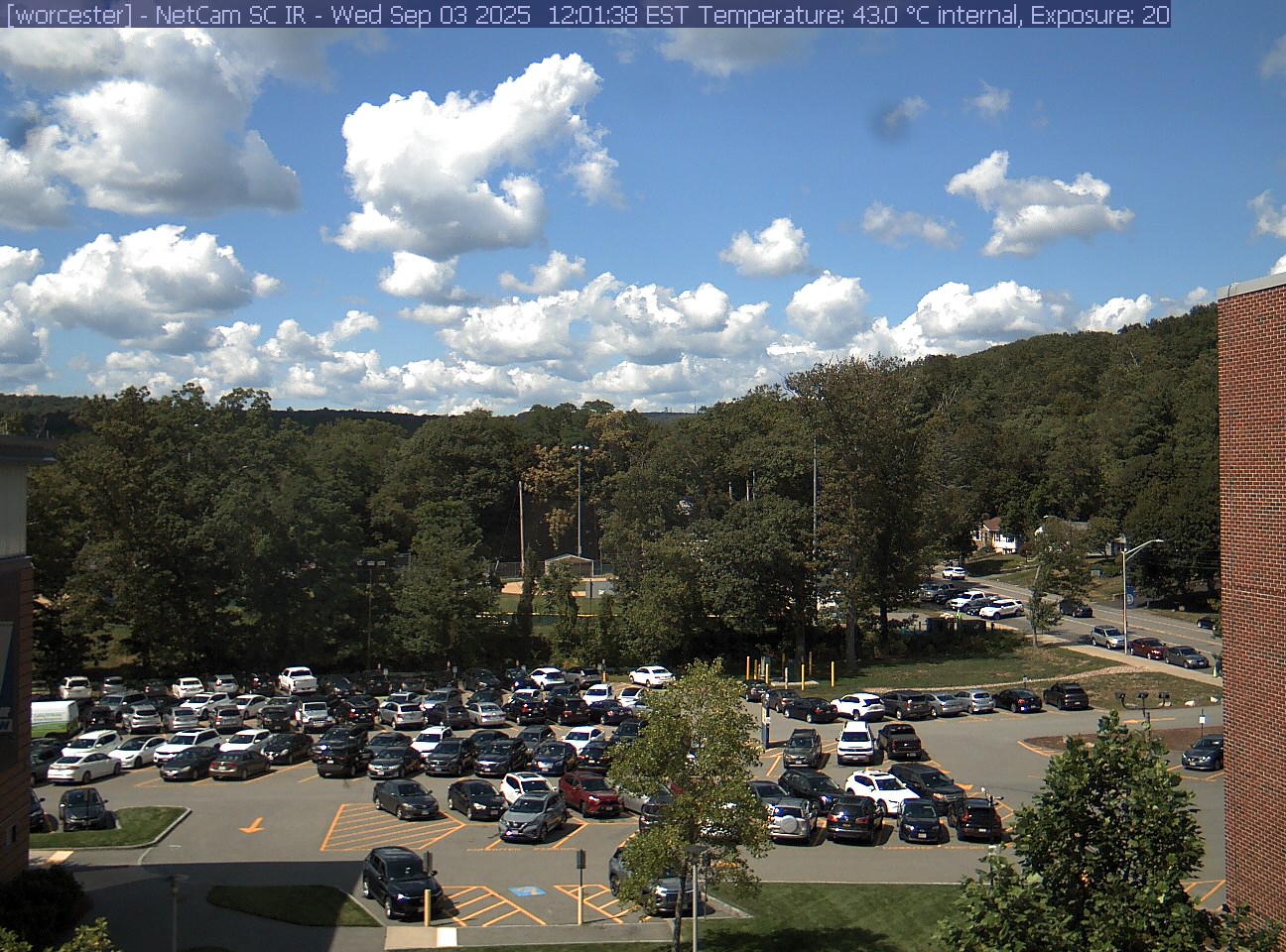 A wooded hill rises beyond a campus car park and a ball field, with a brick wall at the right edge. Cars, buildings and sky take up much of the view.
