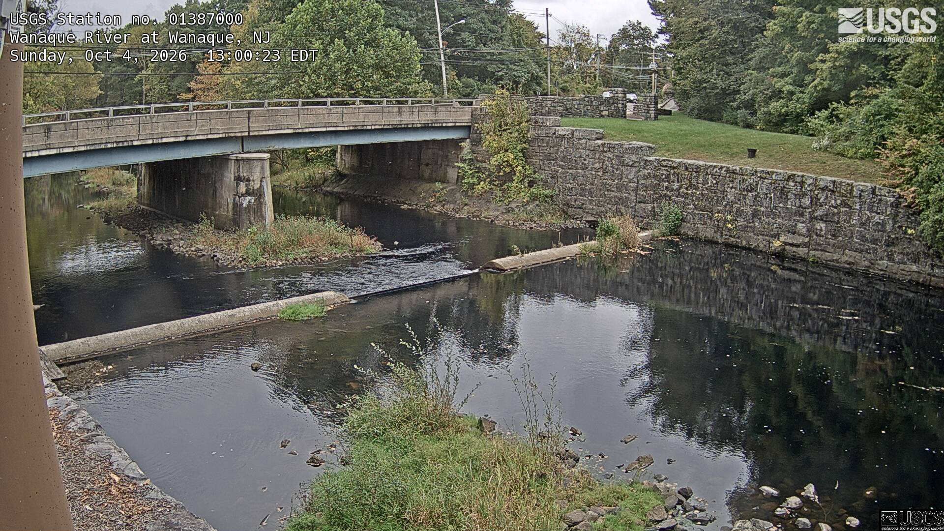 Clear trees around a bridge and stone-walled riverbank, with mostly green crowns and some yellow/orange foliage. The water and structures take most of the lower frame; trees across the upper portion are legible.