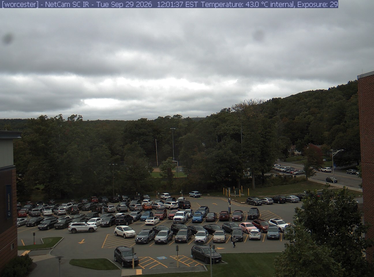 A wooded hill rises beyond a campus car park and a ball field, with a brick wall at the right edge. Cars, buildings and sky take up much of the view.