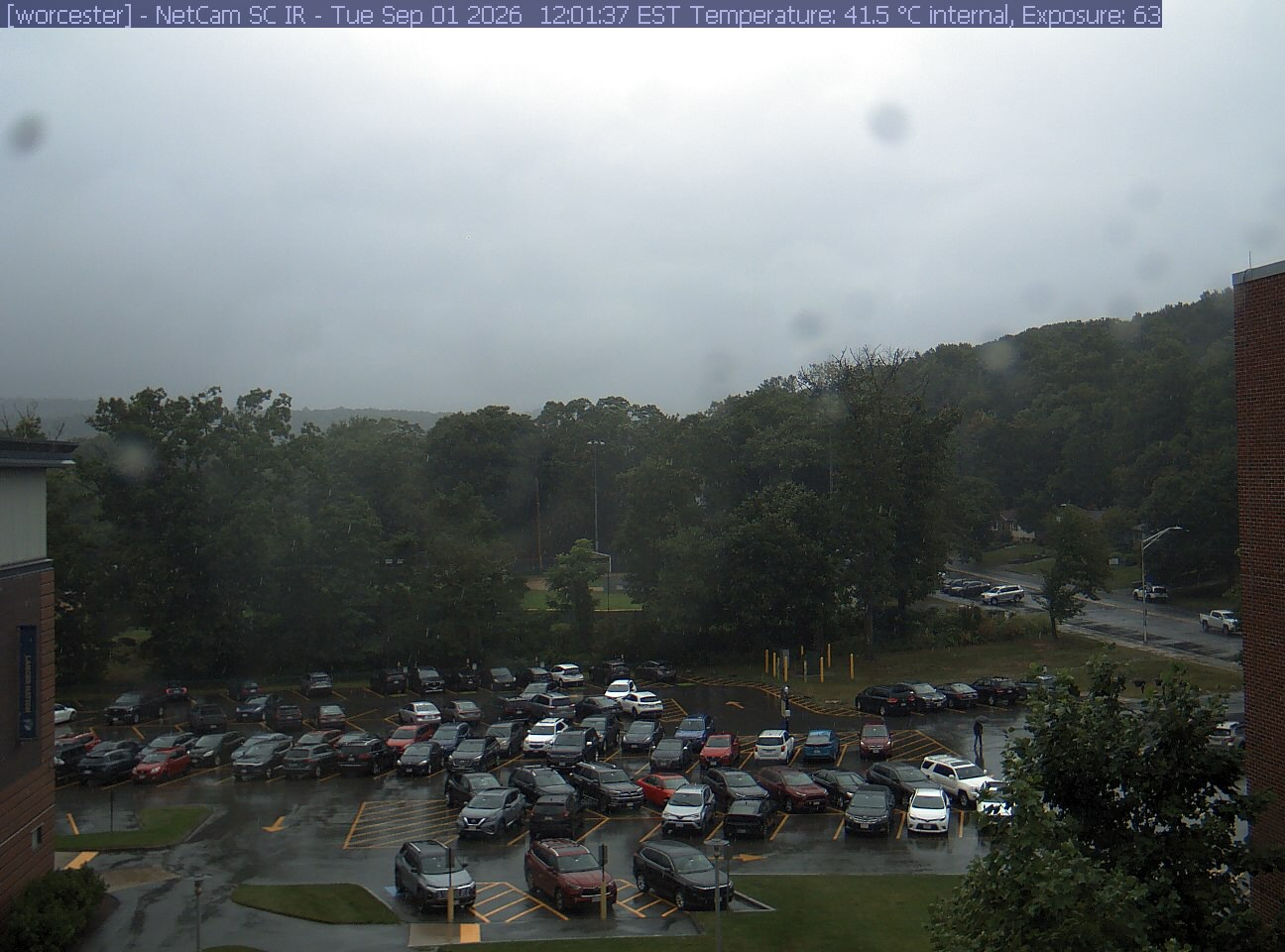 A wooded hill rises beyond a campus car park and a ball field, with a brick wall at the right edge. Cars, buildings and sky take up much of the view.