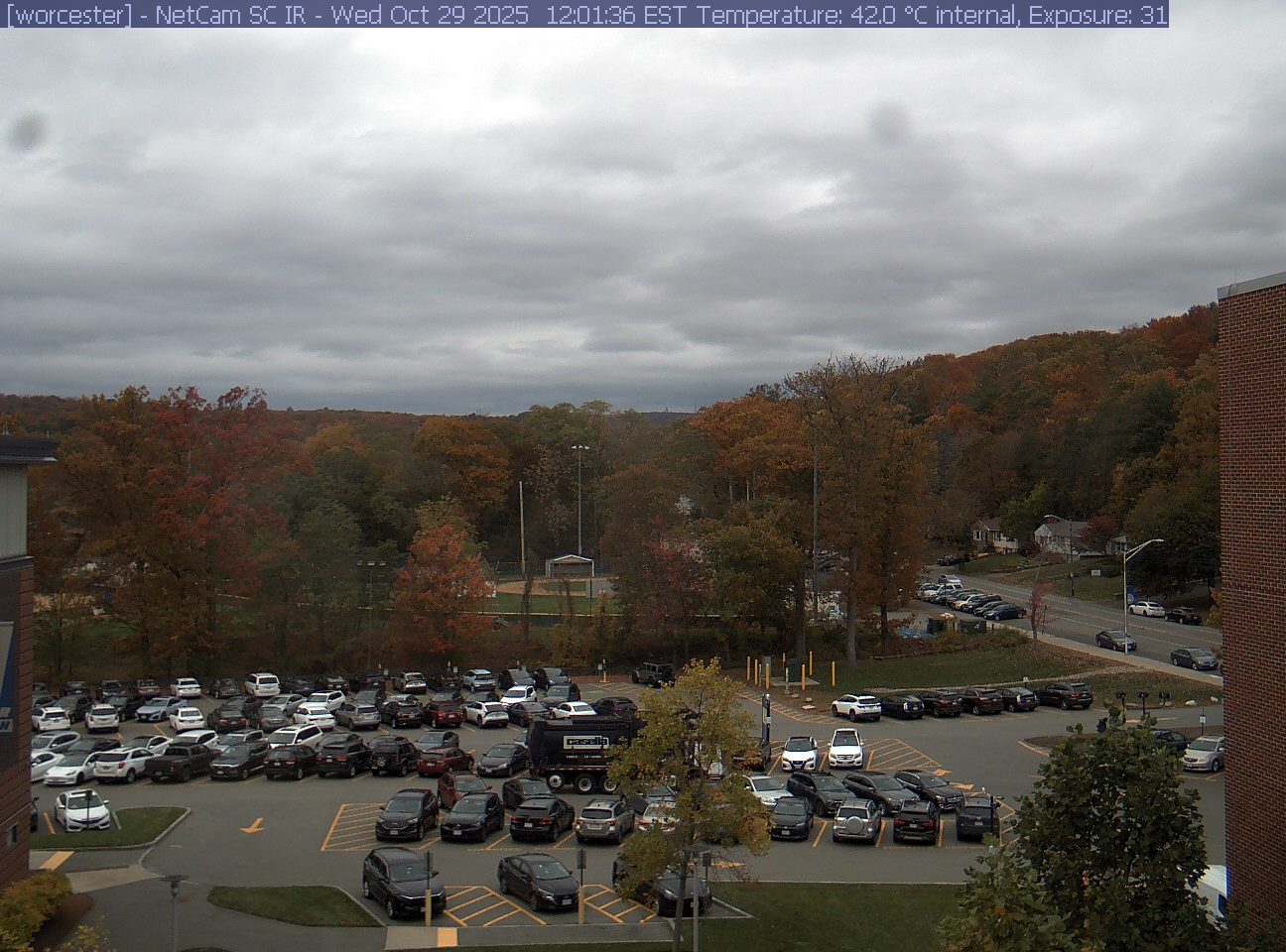A wooded hill rises beyond a campus car park and a ball field, with a brick wall at the right edge. Cars, buildings and sky take up much of the view.