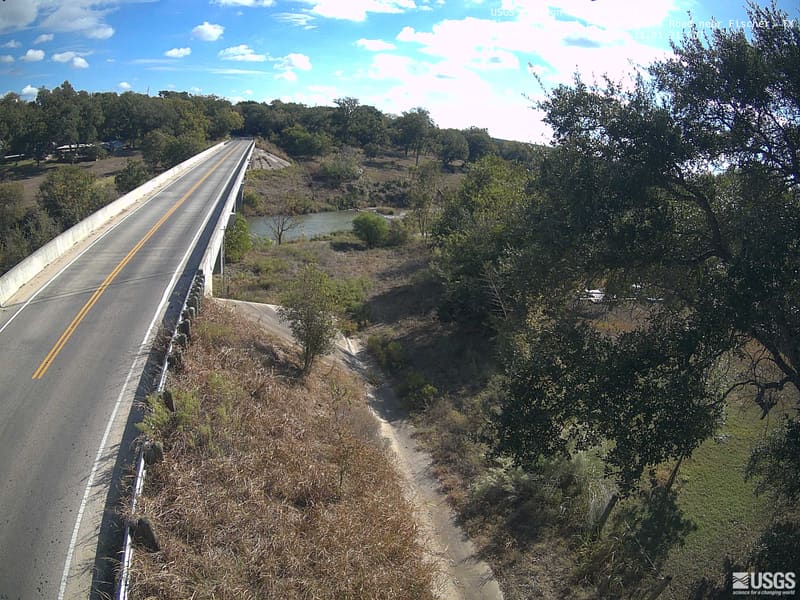 A road bridge crosses the Blanco River valley, with live oaks and scrub along the banks under a wide sky.