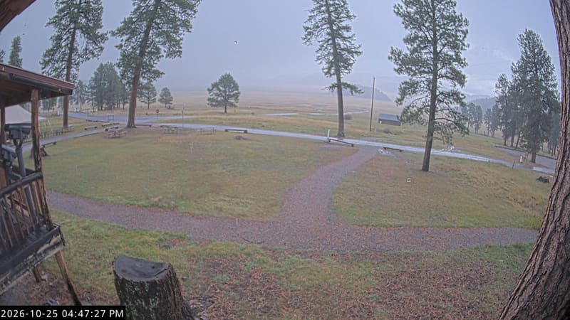 The wide grassland of the Valle Grande beyond a cabin porch and a gravel path, with ponderosa pines and the caldera’s wooded rim in the distance.