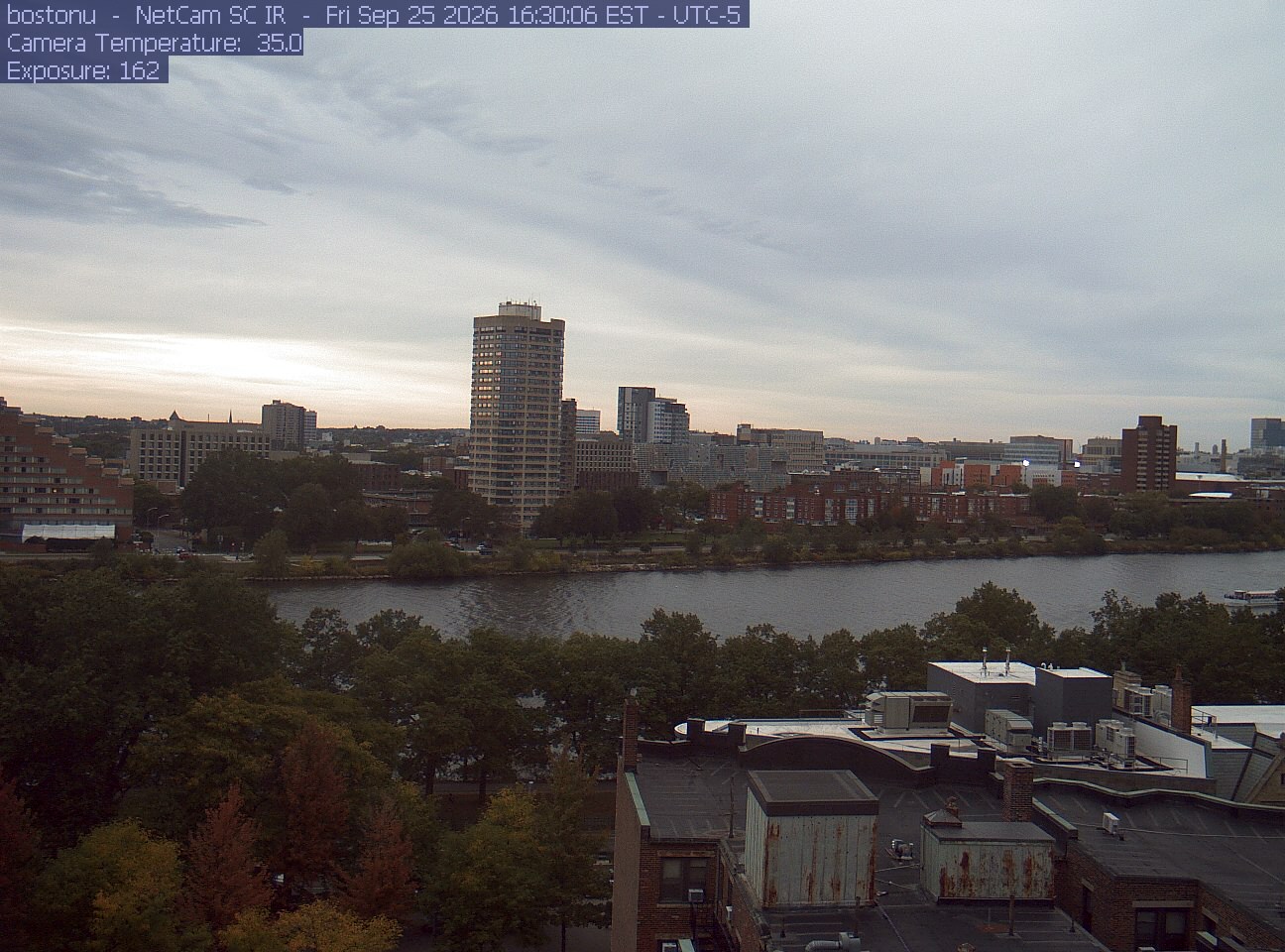 Trees line the river below the city skyline, with a roof edge in the foreground.