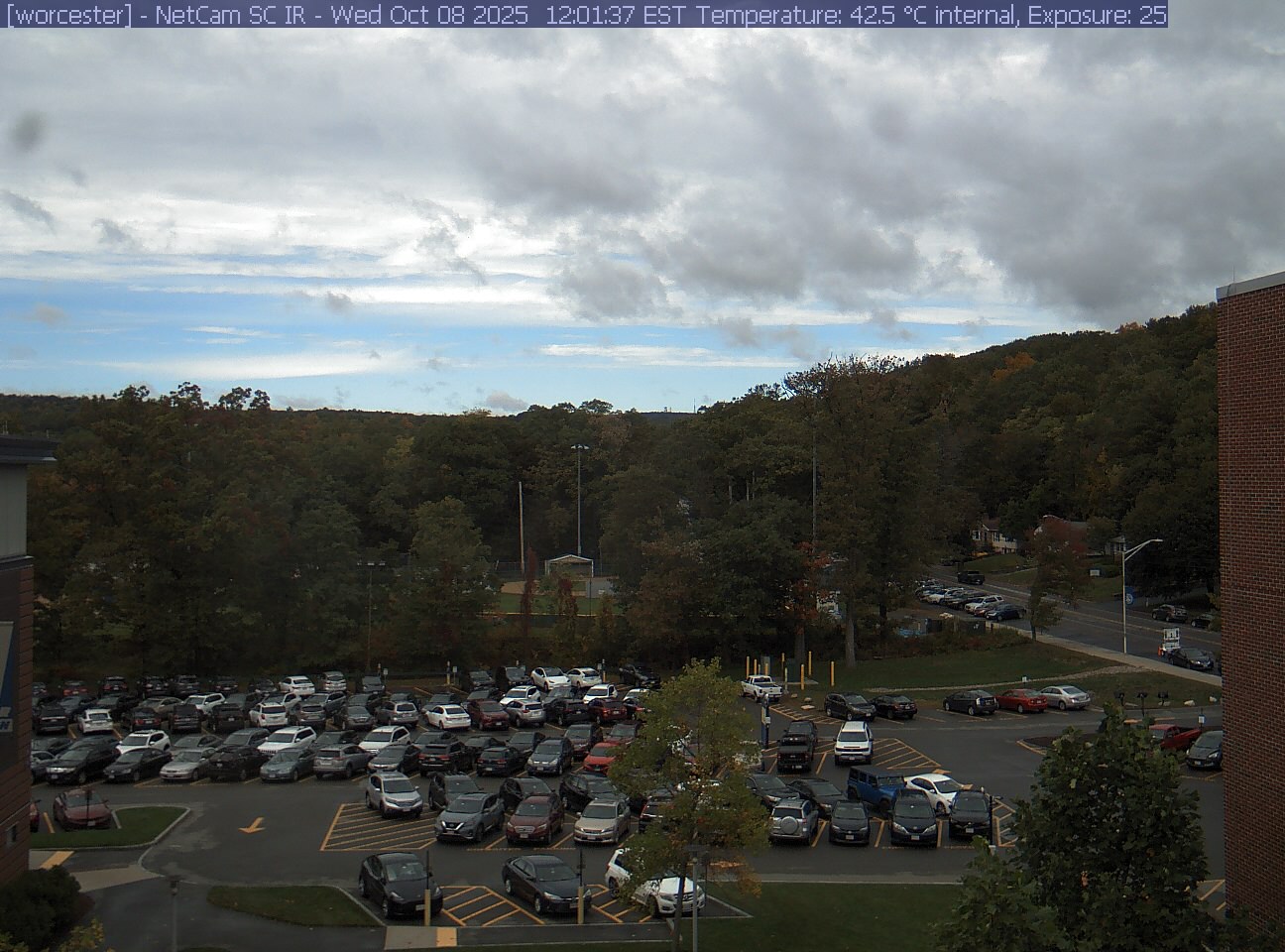 A wooded hill rises beyond a campus car park and a ball field, with a brick wall at the right edge. Cars, buildings and sky take up much of the view.