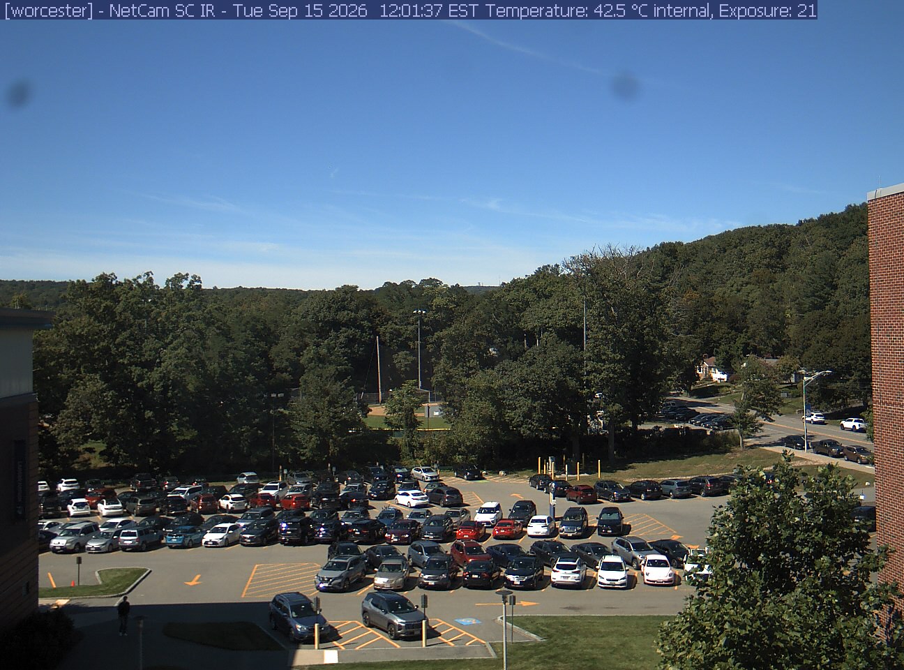 A wooded hill rises beyond a campus car park and a ball field, with a brick wall at the right edge. Cars, buildings and sky take up much of the view.