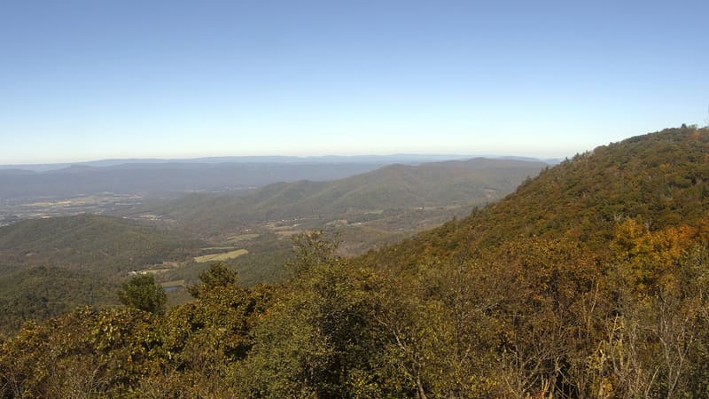 A wooded slope in the near foreground still mostly green with olive and gold crowns, the Shenandoah Valley’s fields and a small pond below, and ridge after ridge fading to blue beyond.