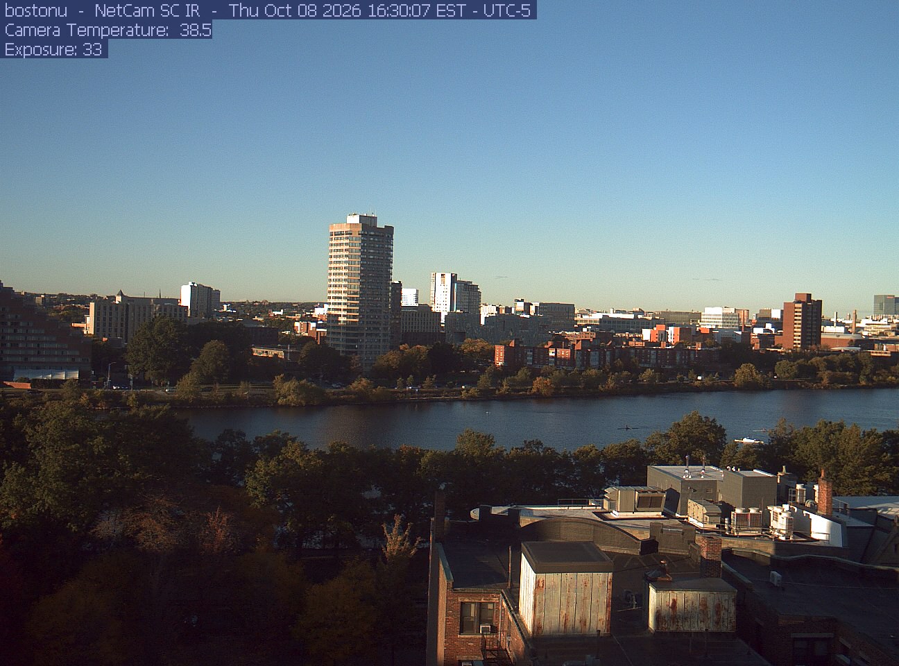 Trees line the river below the city skyline, with a roof edge in the foreground.
