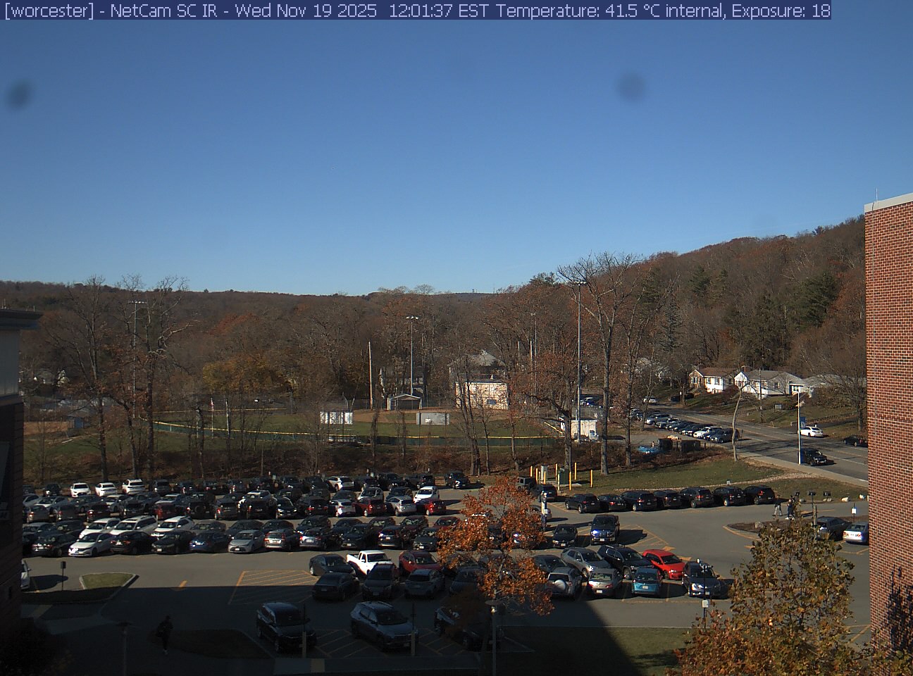 A wooded hill rises beyond a campus car park and a ball field, with a brick wall at the right edge. Cars, buildings and sky take up much of the view.