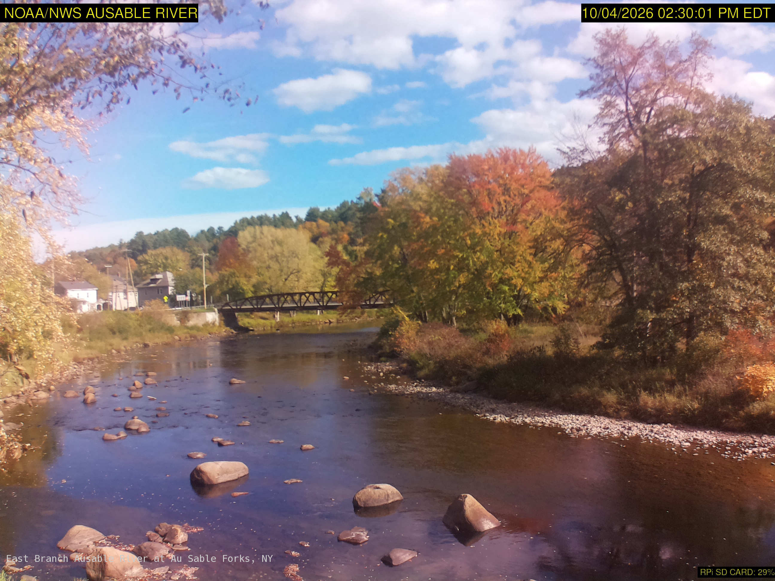 A broad rocky river with a footbridge and large yellow, orange, red and green tree crowns along both banks. Some softness remains, but broad crown color and the riverside setting are clear. The original credits NOAA/NWS; its clock is 15 minutes earlier than the USGS listing, which is a delivery timestamp here.