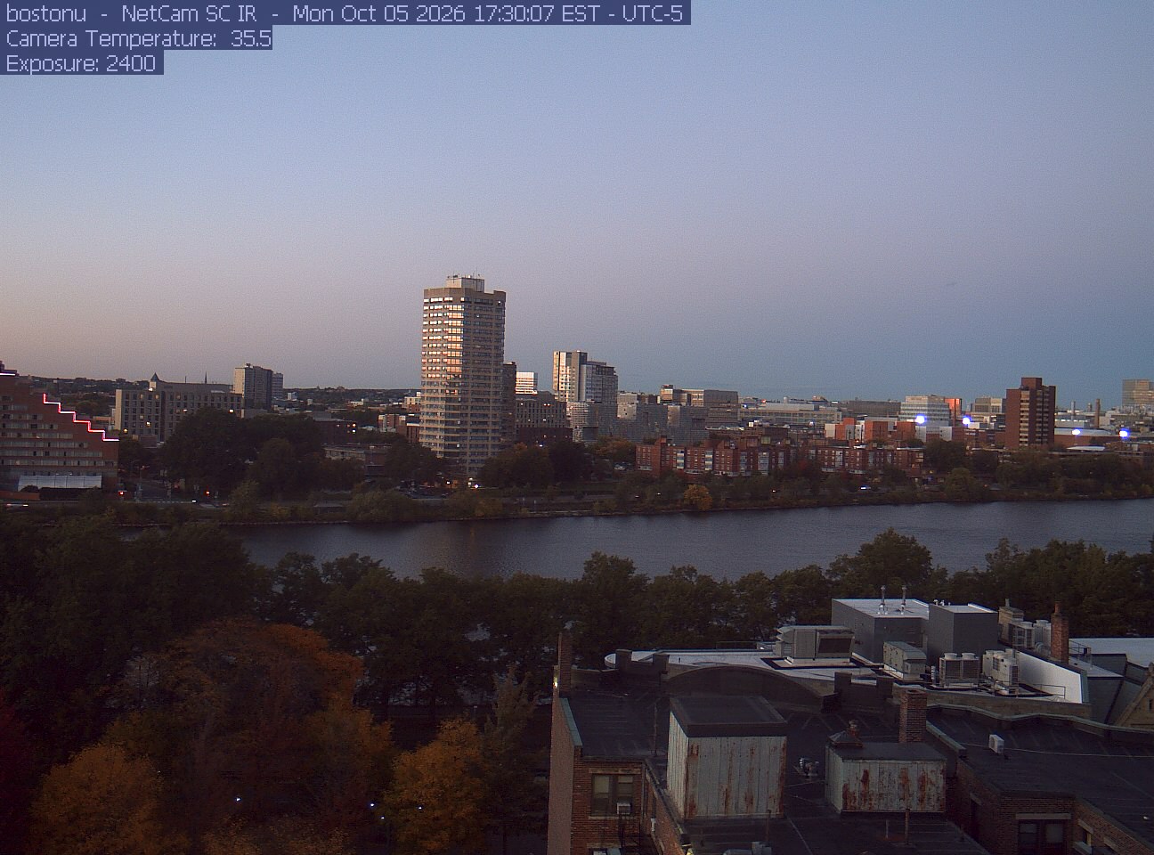 Trees line the river below the city skyline, with a roof edge in the foreground.