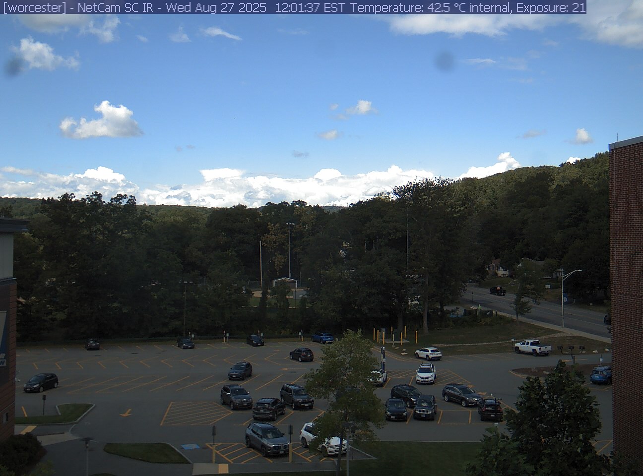 A wooded hill rises beyond a campus car park and a ball field, with a brick wall at the right edge. Cars, buildings and sky take up much of the view.