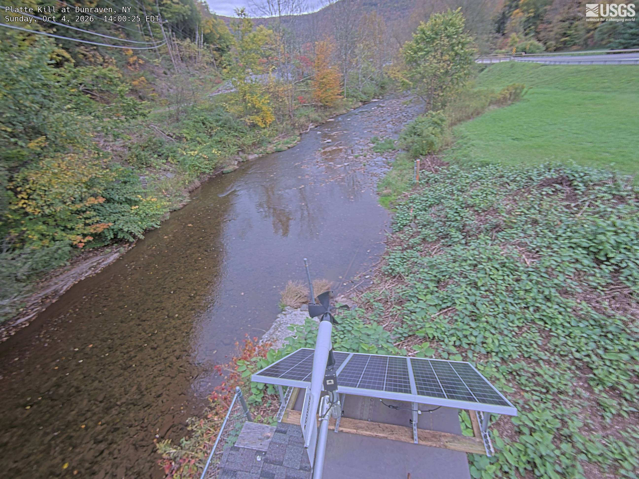 A long rocky creek runs through a small valley, bordered by green, yellow and orange trees with a wooded slope behind.