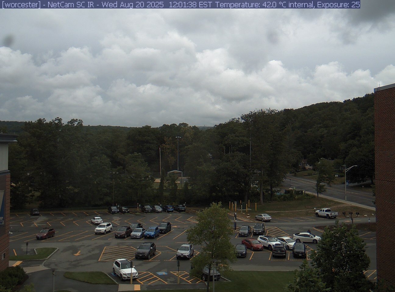 A wooded hill rises beyond a campus car park and a ball field, with a brick wall at the right edge. Cars, buildings and sky take up much of the view.