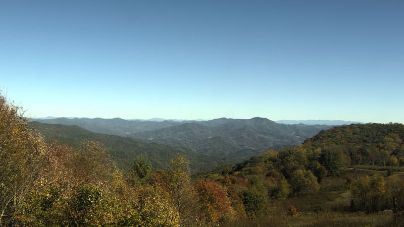 Red, orange and gold crowns in the near foreground and a wide hillside of green turning yellow and rust, with the Smokies’ ridges going blue beyond under a line of fair-weather cloud.
