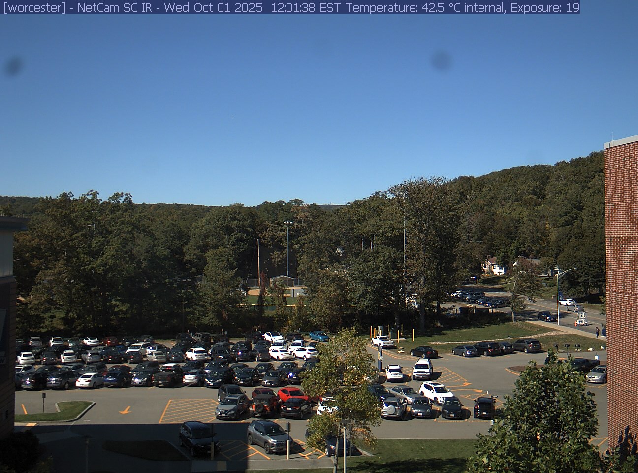 A wooded hill rises beyond a campus car park and a ball field, with a brick wall at the right edge. Cars, buildings and sky take up much of the view.