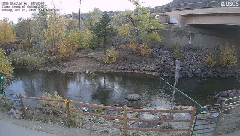 Clear Creek runs past a footbridge railing, with golden cottonwoods and a road bridge beyond.