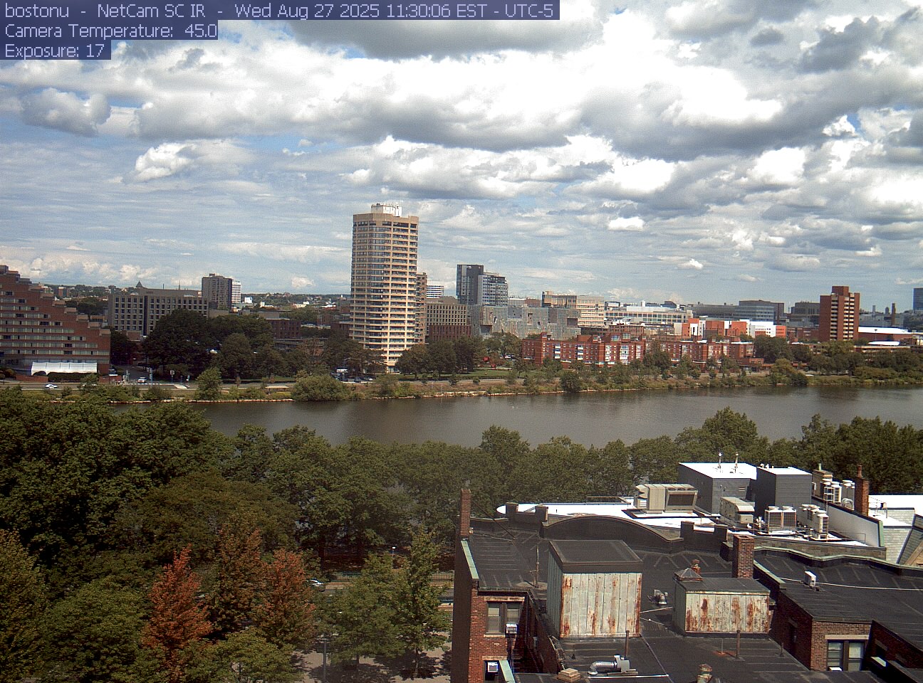 Trees line the river below the city skyline, with a roof edge in the foreground.