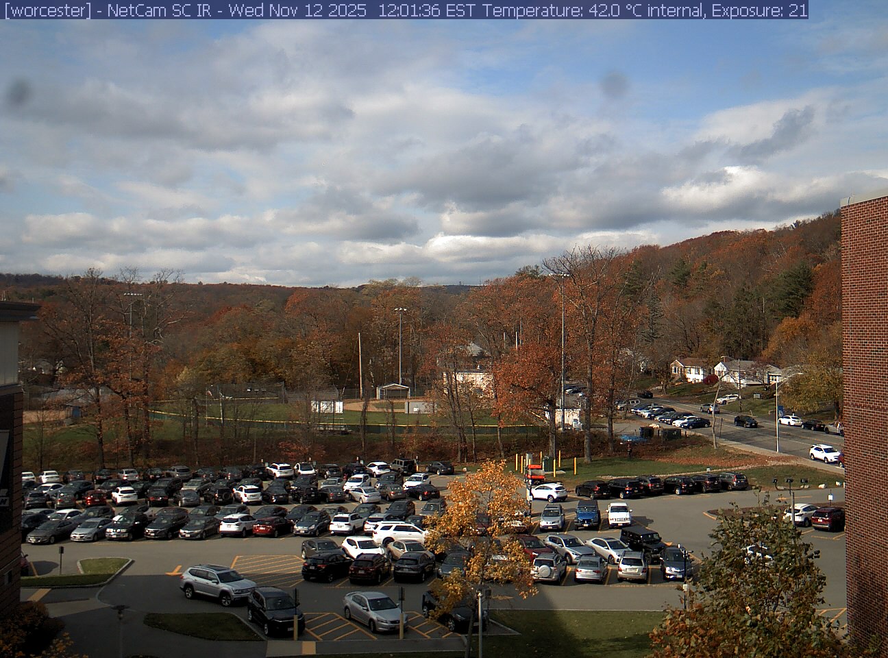 A wooded hill rises beyond a campus car park and a ball field, with a brick wall at the right edge. Cars, buildings and sky take up much of the view.
