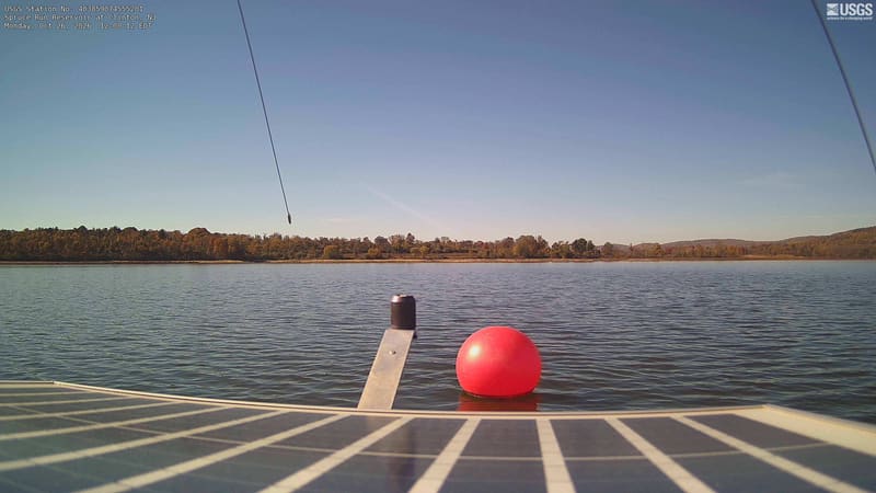 Low wooded hills in gold, rust and muted green stretch across the reservoir beneath a wide sky. A solar panel, red buoy and hanging cables sit close to the camera, with the autumn shore beyond.
