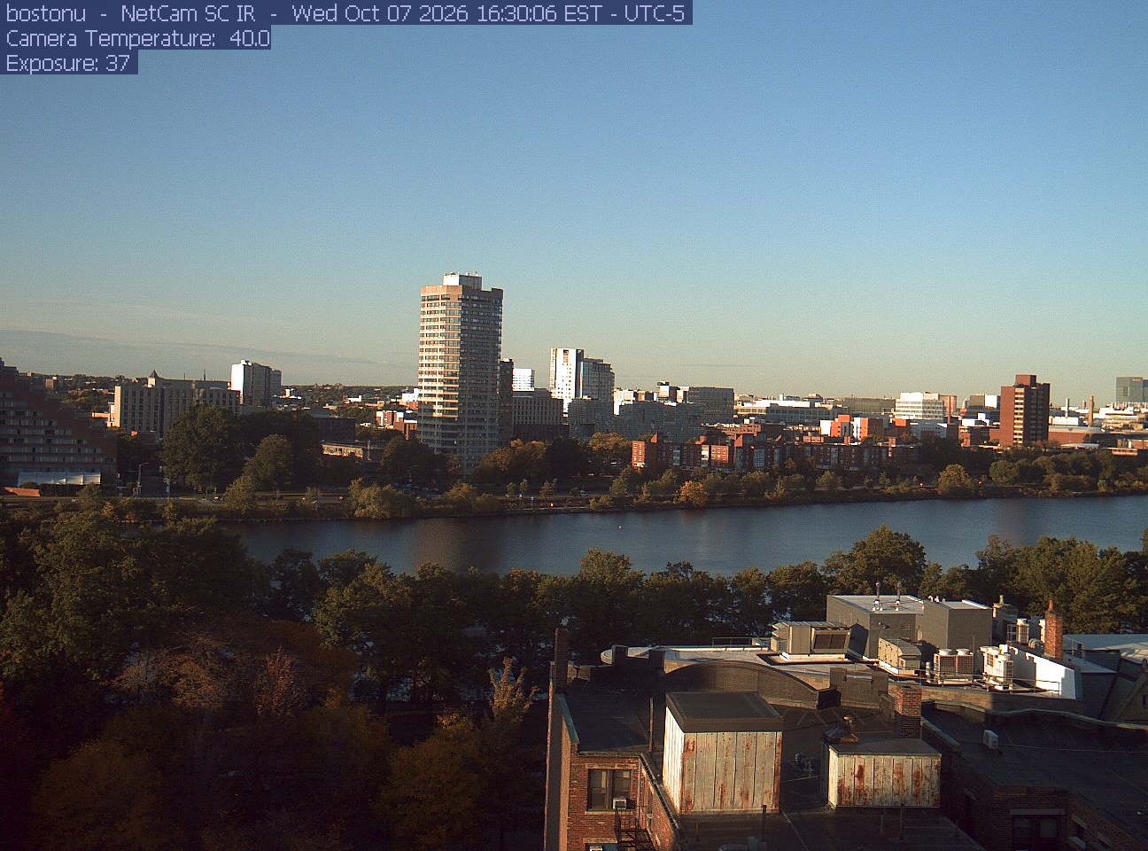Trees line the river below the city skyline, with a roof edge in the foreground.
