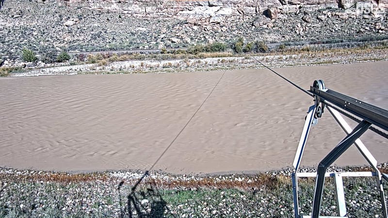 The broad brown river passes below a rocky slope, with low green and muted yellow shrubs along its banks. A metal arm reaches in from the right, with the hills visible beyond.