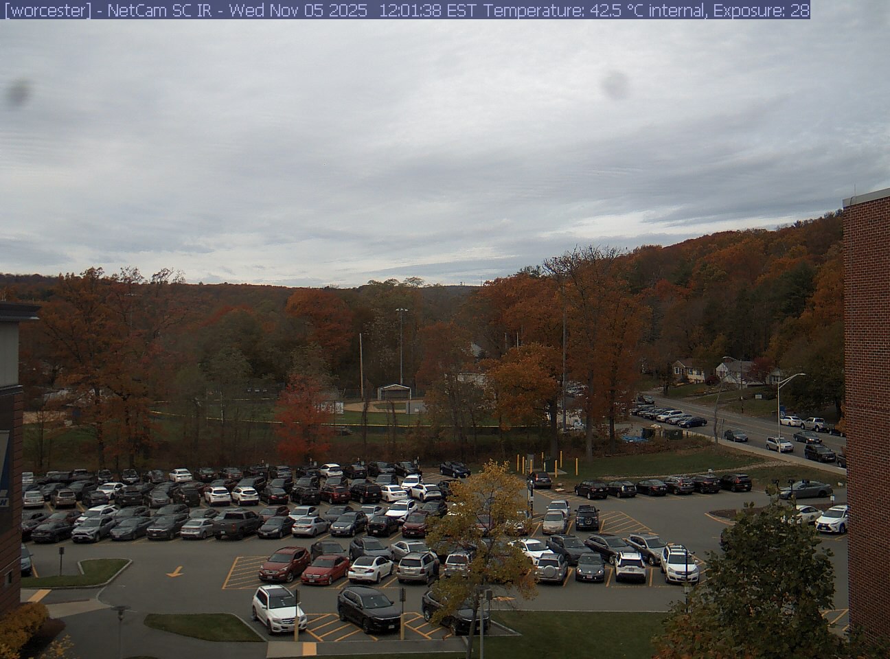 A wooded hill rises beyond a campus car park and a ball field, with a brick wall at the right edge. Cars, buildings and sky take up much of the view.