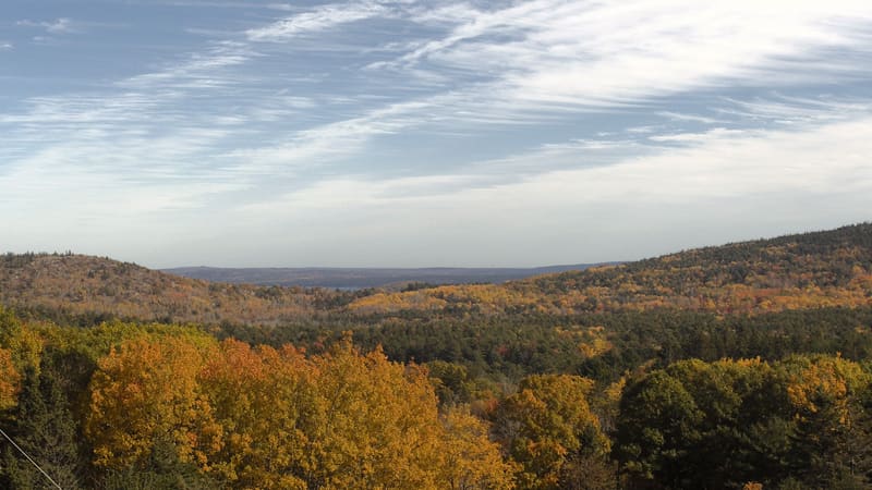 Gold and orange crowns in the near foreground, a slope of yellow and red hardwoods among dark pines rising to a bare granite hilltop at the left, and the bay beyond under a clear sky.