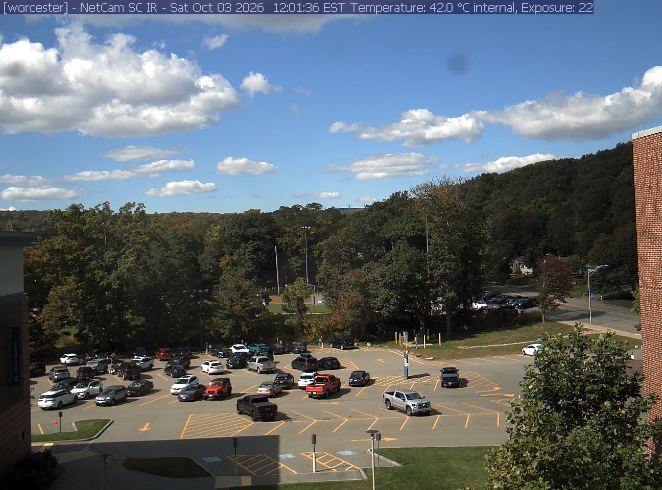 A wooded hill rises beyond a campus car park and a ball field, with a brick wall at the right edge. Cars, buildings and sky take up much of the view.