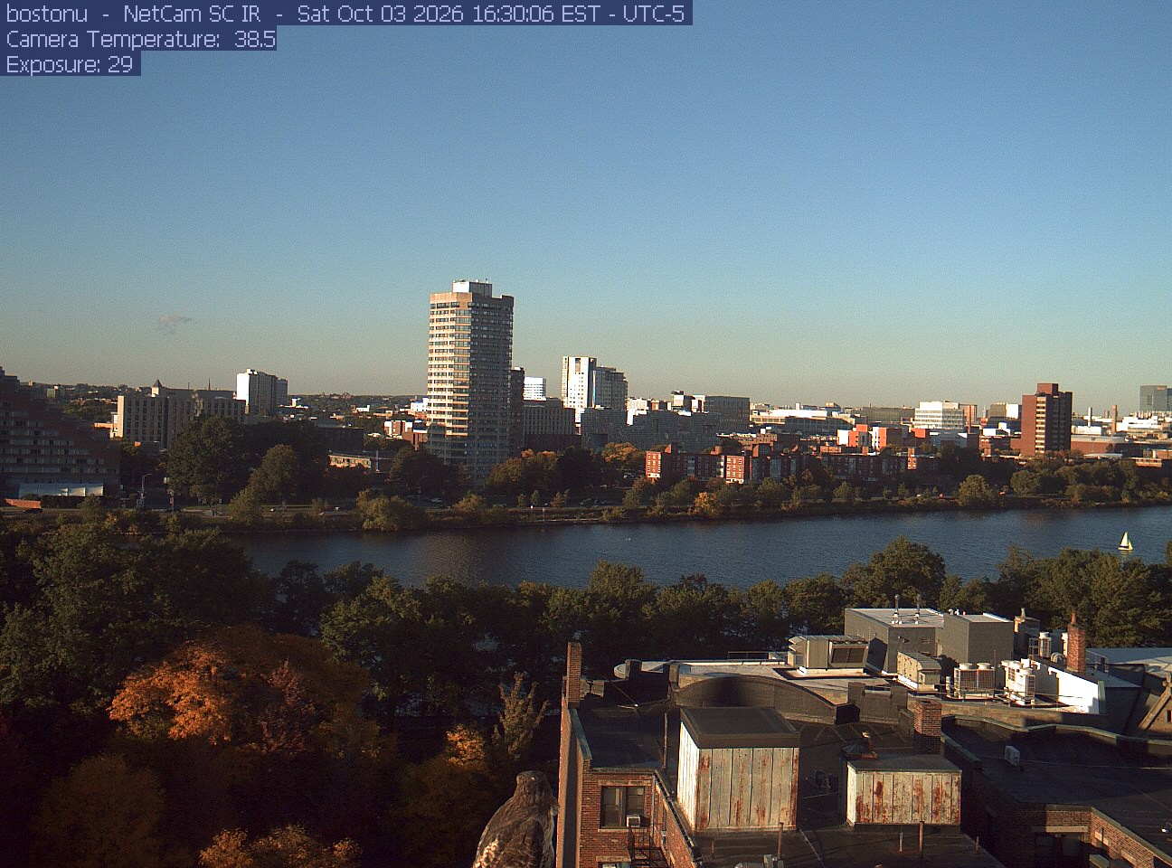Trees line the river below the city skyline, with a roof edge in the foreground.