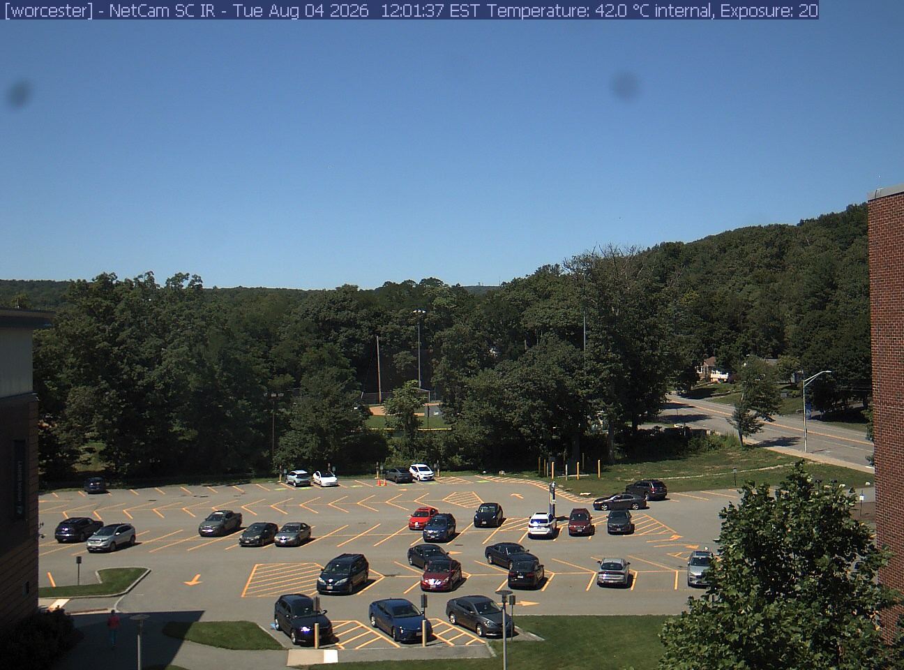 A wooded hill rises beyond a campus car park and a ball field, with a brick wall at the right edge. Cars, buildings and sky take up much of the view.