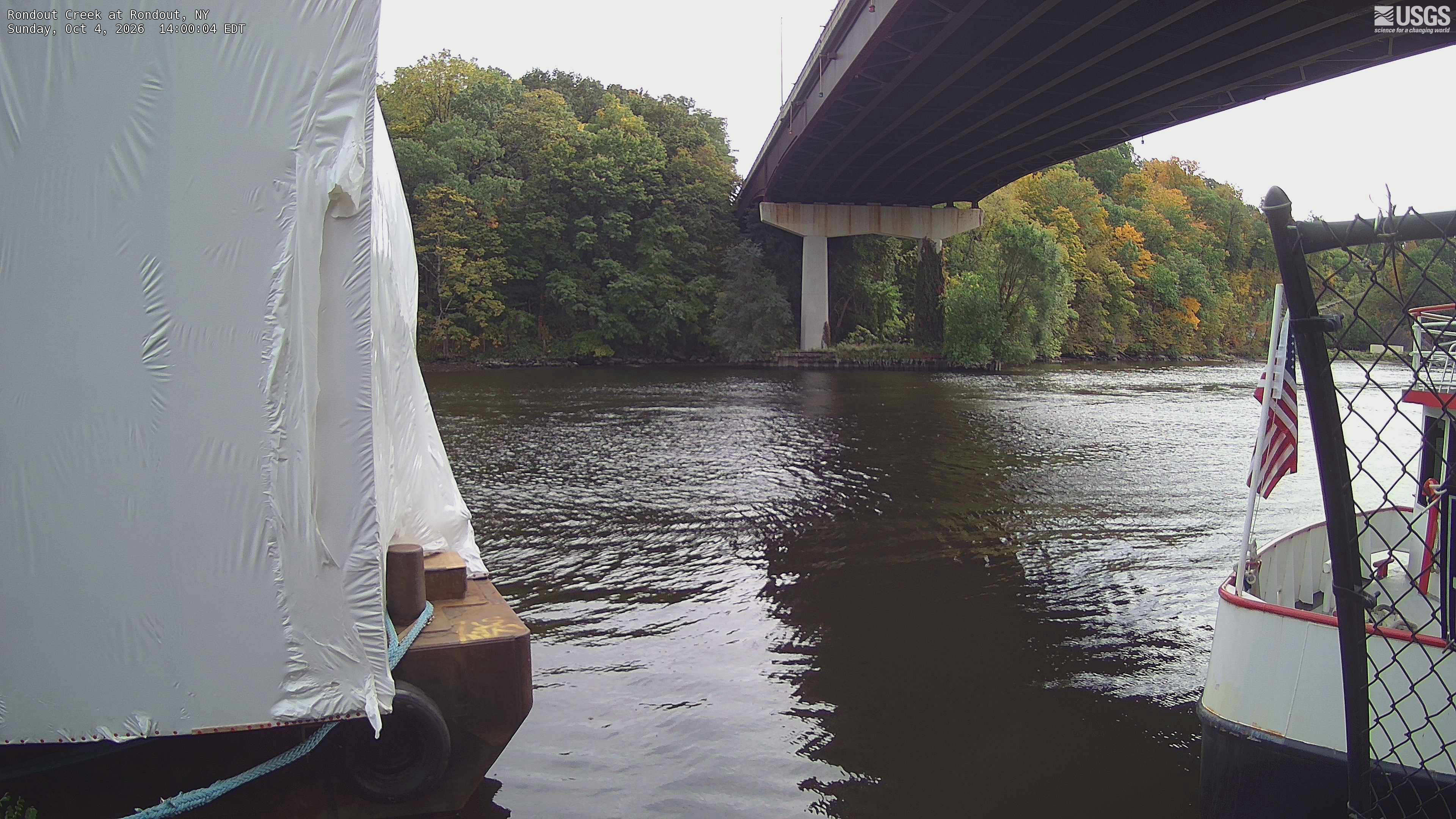 A clearly resolved wooded bank across the creek, with green, yellow and orange crowns beside a large road bridge. A covered boat obscures the left third, with another boat and fence at right.