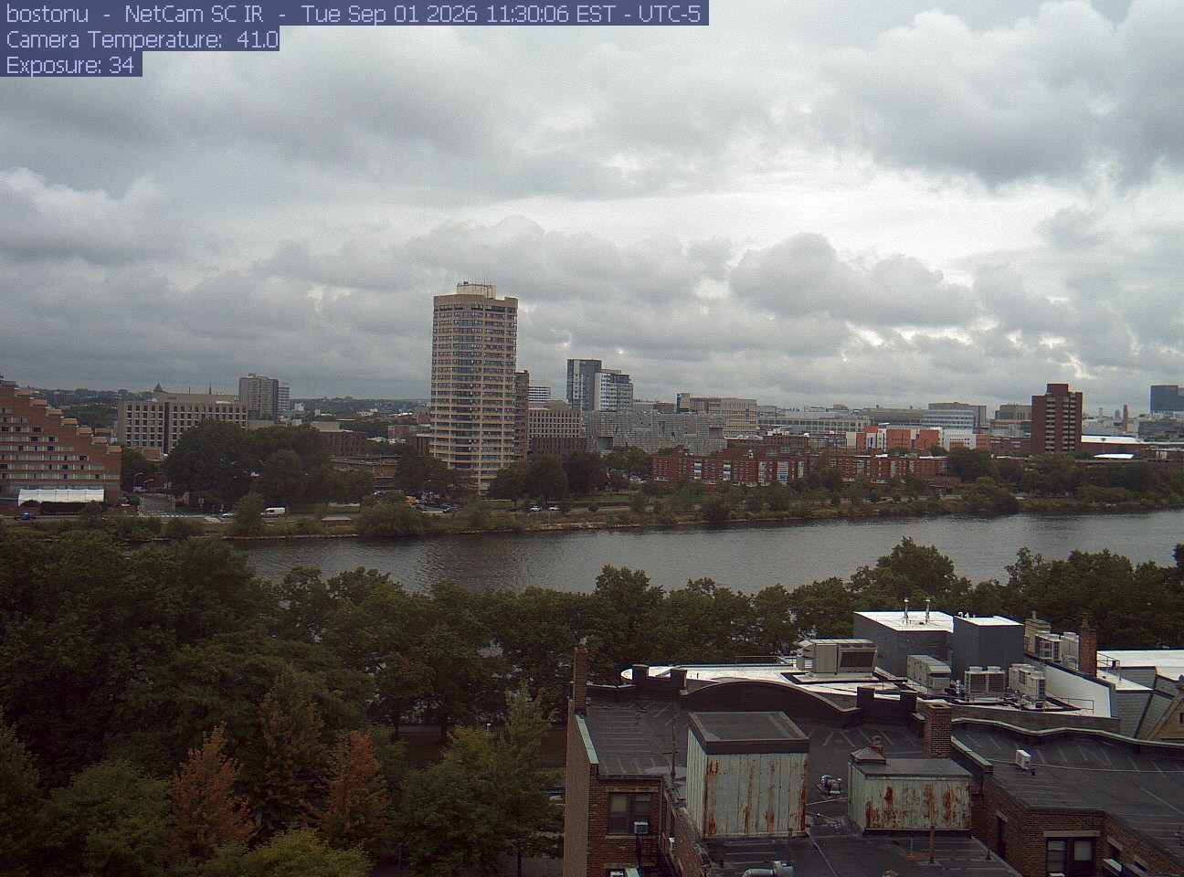 Trees line the river below the city skyline, with a roof edge in the foreground.